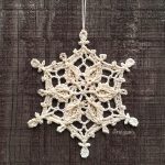 A cream-colored crocheted Winter Realm Snowflake ornament hangs against a dark wooden background.