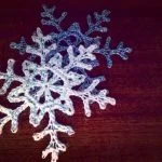 Snowflake #1, a delicate crocheted decoration, lies alongside a light blue counterpart on a dark wooden surface, their lacy edges overlapping gently.