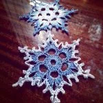 Two intricately crocheted snowflake ornaments, including Snowflake #2 in blue, are placed on a polished wooden surface with blurred lights in the background.