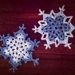 Two intricately crocheted snowflake ornaments, including Snowflake #3 in blue and another in white, are displayed on a dark wooden surface.