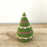 A small, cone-shaped crochet Tiny Lights Christmas Tree with pink and white decorations and a gold bead on top, placed on a wooden surface against a plain wall.