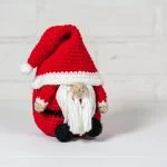 A small crochet Santa Claus Gnome with a red hat, white beard, and black feet sits on a white surface against a light brick background.