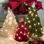 Three crocheted Christmas trees from the Bobble Stitch Christmas Tree Trio, one white, one green, and one red with lights, are displayed on a table amid festive decorations and red poinsettias in the background.