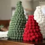 The Bobble Stitch Christmas Tree Trio in green, red, and white yarn is displayed on a wooden surface, with blurred holiday decorations in the background.