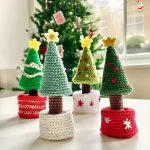 Four crocheted Christmas tree decorations with festive embellishments are displayed in front of a decorated Christmas Tree with Pot near a window.