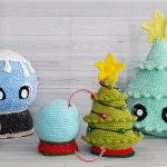 Three crocheted holiday decorations: a 3-in-1 Reversible Christmas Tree with ornaments and a star, a blue snow globe base, and a turquoise tree with a yellow star, all displayed on a wooden surface.
