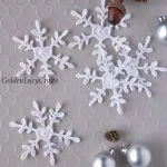 Three crocheted white heart snowflake ornaments are displayed on a light hexagonal surface, with silver baubles and pinecone decorations nearby.