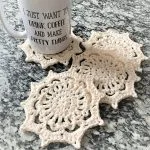 A white mug with black text sits on a crocheted cream Zeta Snowflake coaster, with two more matching coasters on a marbled countertop.
