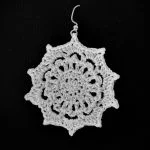 A single, intricately crocheted earring in a circular, floral pattern—reminiscent of a Zeta Snowflake—is displayed against a solid black background.