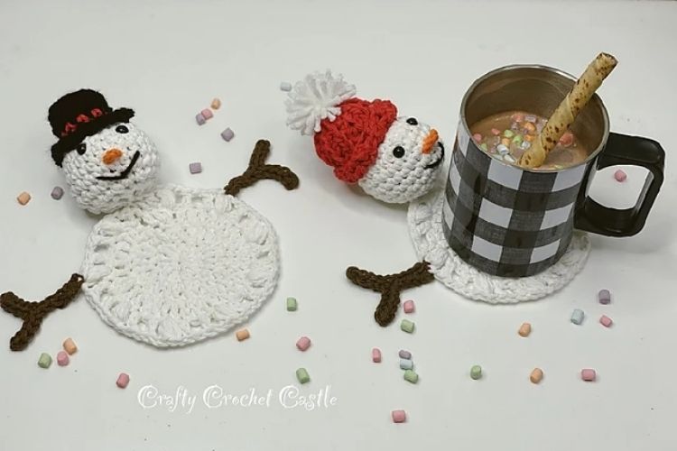 Two crocheted snowmen sitting next to a cup of hot cocoa.