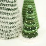 Three crocheted Cone Christmas Trees in green and white yarn, adorned with pearl-like beads and tinsel accents, stand elegantly on a white surface.