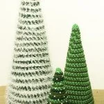 Three cone-shaped crochet Christmas trees, including two green and one adorned with white tinsel, are displayed on a burlap surface, showcasing charming Cone Christmas Trees against a plain background.