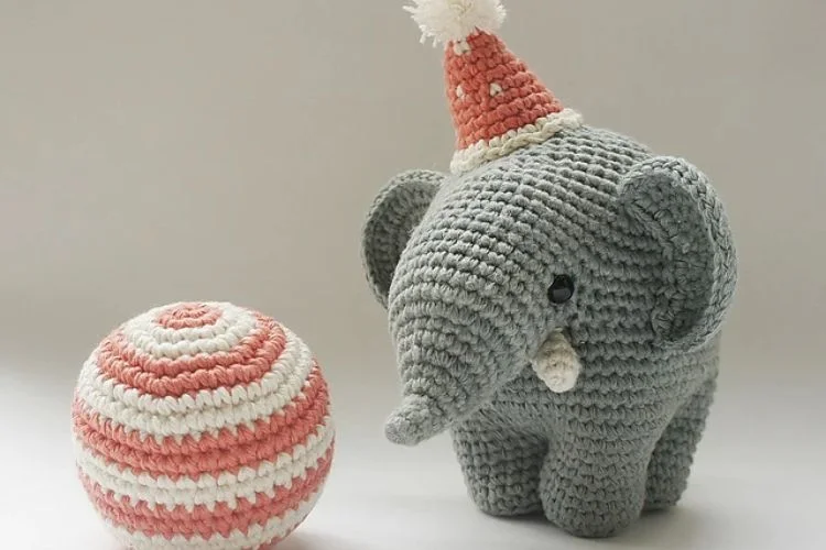 A crocheted elephant with a birthday hat next to a ball.