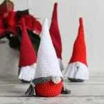 Four crocheted Scandinavian Christmas Gnome figures with long hats and beards are displayed on a light surface, with a poinsettia plant in the background.