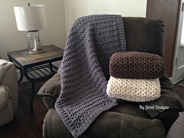 A brown recliner with a gray Jenni's Favorite Chunky Throw Blanket draped over it and two folded crocheted blankets, one brown and one cream, stacked on the seat; a side table and lamp are nearby.