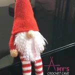 Crocheted Scandinavian Christmas Gnome doll with a tall red hat, striped red and white legs, a white beard, and black shoes, sitting on a dark surface. 