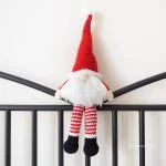 A Scandinavian Santa Gnome doll with a tall red hat, white beard, and striped red-and-white legs sits on a black metal railing against a white wall.