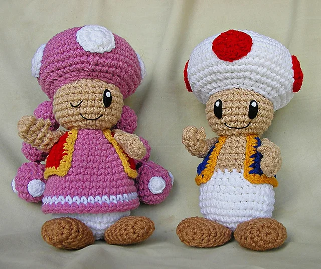 Two crocheted dolls resembling Toad and Toadette from Mario games—one with a pink outfit and mushroom cap, the other with a white cap with red spots and blue vest—both smiling.