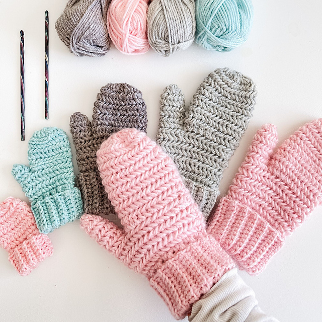Several pairs of handmade Heavenly Herringbone Mittens in pastel colors are displayed alongside yarn balls and crochet hooks on a white surface.