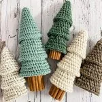 Four crocheted Christmas tree ornaments with cinnamon stick trunks, inspired by Rustic Farmhouse Tiered Trees, are arranged on a white wooden surface in shades of green, cream, and brown.