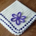 a white crochet baby blanket with a purple flower motif