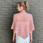 A woman with blonde hair in a ponytail wears the Bella Rosa Shawl—a light pink crocheted piece—over a white outfit, standing in front of a gray brick wall.