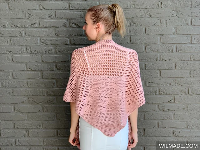 A woman with blonde hair in a ponytail wears the Bella Rosa Shawl—a light pink crocheted piece—over a white outfit, standing in front of a gray brick wall.