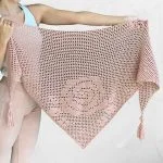A person holds up a large, light pink, crocheted Bella Rosa Shawl with tassels on the two bottom corners.