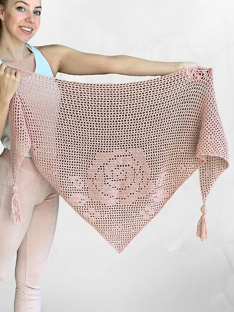 A person holds up a large, light pink, crocheted Bella Rosa Shawl with tassels on the two bottom corners.