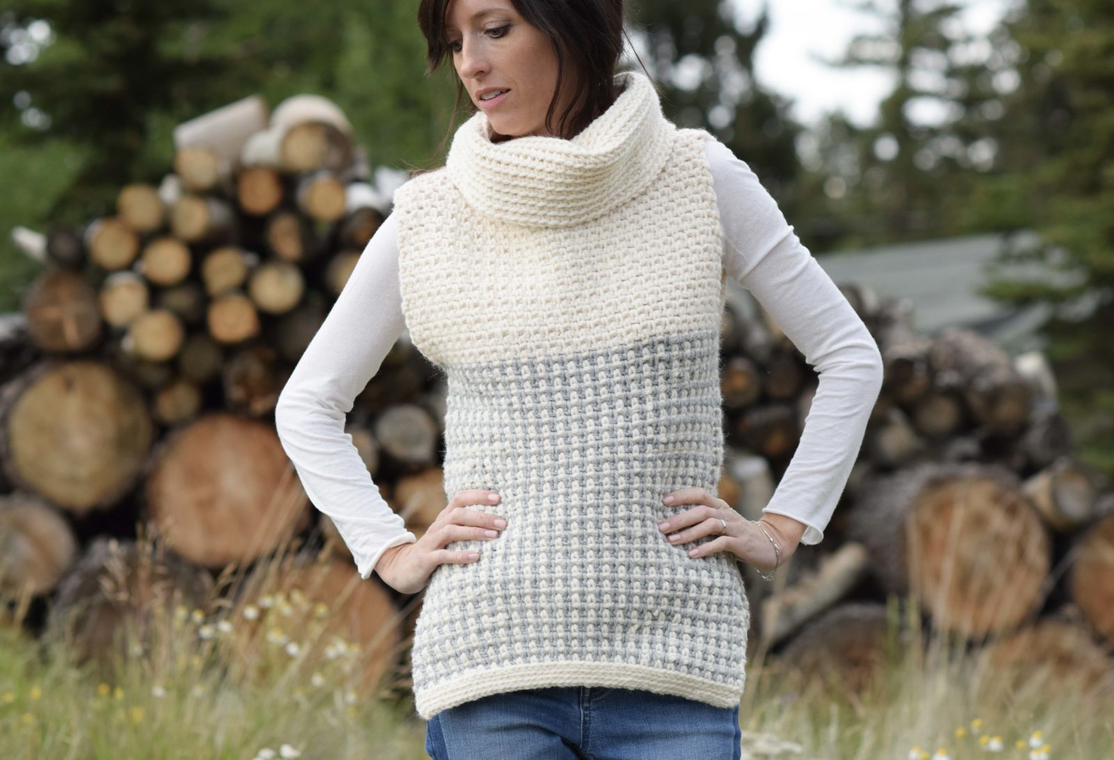 A woman wearing the Easy Cowled Sweater Vest in cream and light gray textured knit stands outdoors in front of a stack of cut logs.