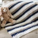 A plush bunny toy rests on a beige couch next to a Done In a Day Baby Blanket, crocheted with white, blue, and brown stripes.