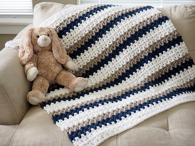 A plush bunny toy rests on a beige couch next to a Done In a Day Baby Blanket, crocheted with white, blue, and brown stripes.