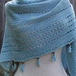 Light blue crocheted shawl with openwork rows, draped over a mannequin, featuring tassels with wooden bead accents along the edge—a breezy S'up Coconut style accessory.