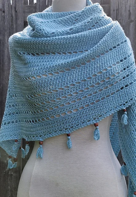 Light blue crocheted shawl with openwork rows, draped over a mannequin, featuring tassels with wooden bead accents along the edge—a breezy S'up Coconut style accessory.