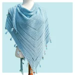 A light blue S'up Coconut knitted shawl with a triangular shape, eyelet pattern, and tassel decorations draped over a mannequin against a pale blue background.
