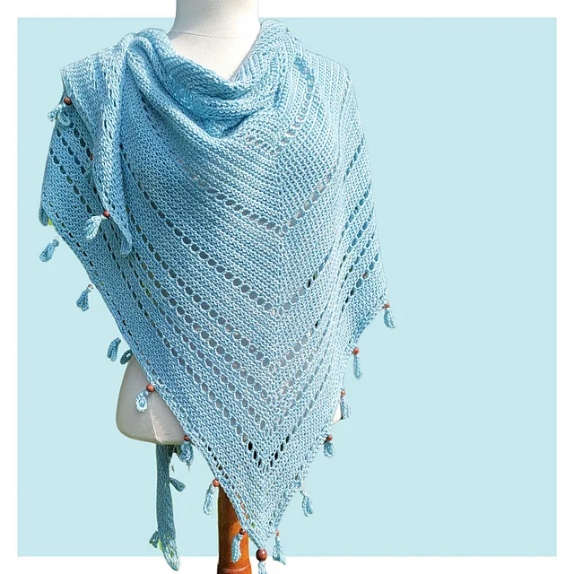 A light blue S'up Coconut knitted shawl with a triangular shape, eyelet pattern, and tassel decorations draped over a mannequin against a pale blue background.