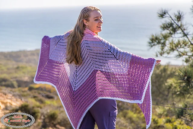 Woman standing outdoors with arms outstretched, wearing a purple and white Tulip Chevron Shawl, with a scenic ocean and trees in the background.