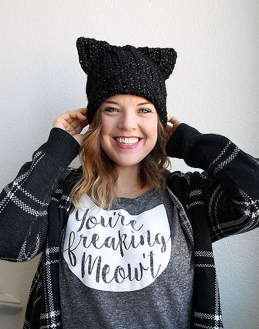 A smiling woman wears a Black Cat Slouch Hat with playful cat ears, a plaid cardigan, and a gray shirt that says 