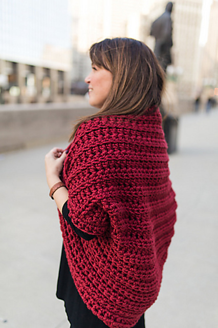 A woman stands outdoors wearing a chunky red crocheted Juno Shrug over a black top, with city buildings in the background.