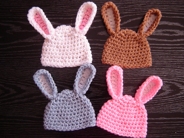 Four Easy Bunny Hats with crocheted bunny ears in white, brown, gray, and pink are arranged on a dark wooden surface.