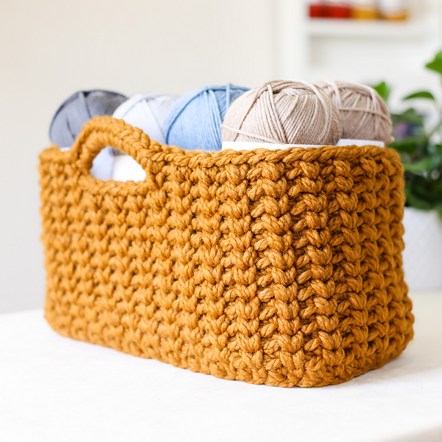 A chunky knit mustard yellow Jessie Stash Basket holds several skeins of yarn in gray, blue, and beige tones.