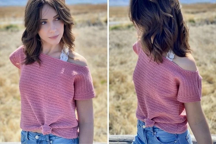 A young woman wearing an off-shoulder pink knitted top and jeans, posing in a field.