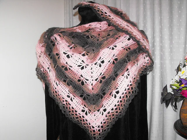 A mannequin draped in a pink and gray Butterfly Stitch Prayer Shawl with lacy, scalloped edges and triangular crochet patterns, set in front of light-colored curtains.