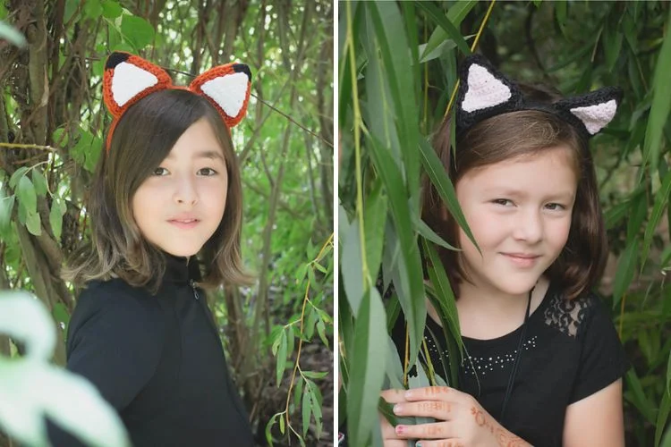 Two young girls wearing animal ear headbands pose playfully among green foliage; one with orange ears, the other with black and white.