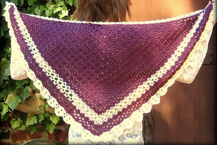 A handmade purple and white crocheted shawl draped over a person's shoulders against a backdrop of green foliage.