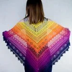 A person stands facing away, displaying a large, triangular Flower Valley Shawl crocheted in a gradient of yellow, orange, red, pink, and purple hues.