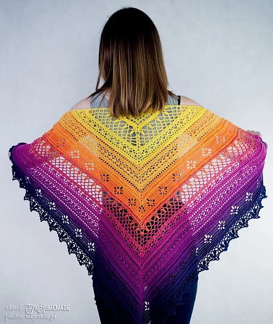 A person stands facing away, displaying a large, triangular Flower Valley Shawl crocheted in a gradient of yellow, orange, red, pink, and purple hues.