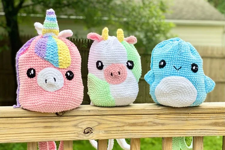 Three crocheted stuffed animals—a pink unicorn, a green and white unicorn, and a blue whale—sitting on a wooden fence with a grassy background.