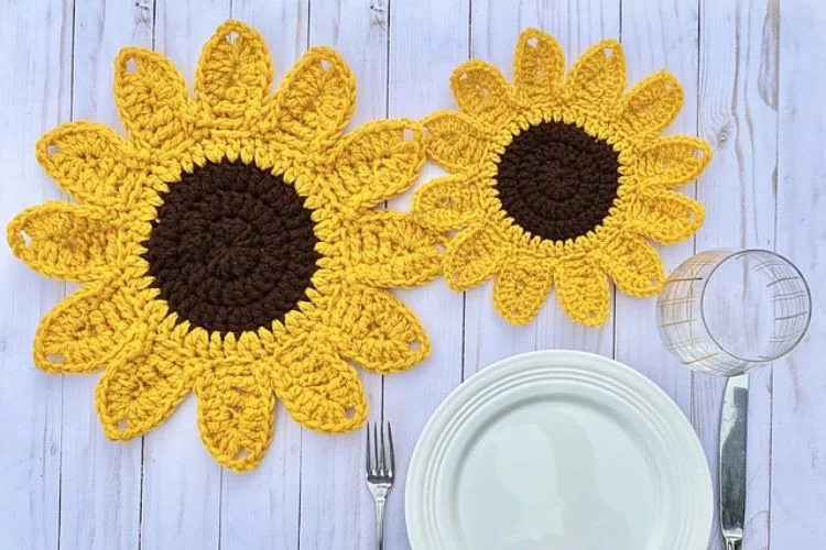 Two crocheted sunflower placemats on a wooden table next to empty dishes and cutlery.