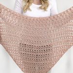 A woman holds up a large, triangular, light pink crocheted shawl with an open, lacy pattern—the perfect Vintage Tulip Shawl for adding timeless elegance to any outfit.
